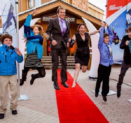 man in suit beams as he walks down a red carpet, with excited, happy, jumping children linking each of his arms and flanking beside them