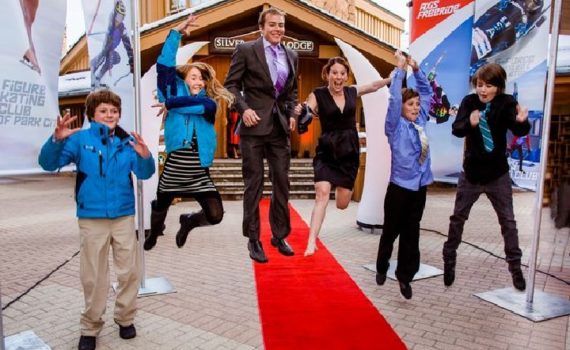 man in suit beams as he walks down a red carpet, with excited, happy, jumping children linking each of his arms and flanking beside them