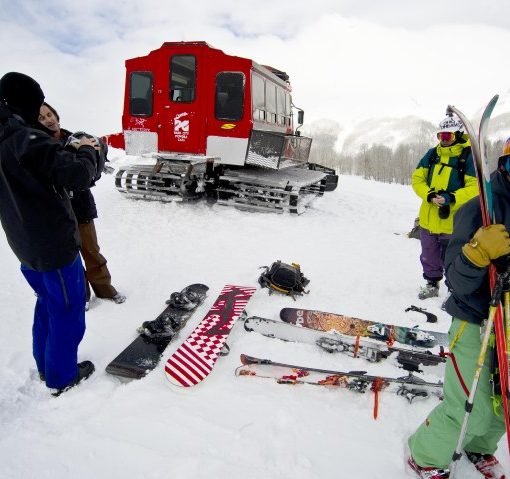 skiers stand near their skis which are laid out on the ground, a powder cat is parked a little ways away