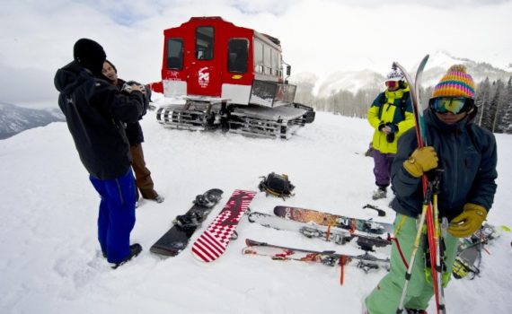 skiers stand near their skis which are laid out on the ground, a powder cat is parked a little ways away