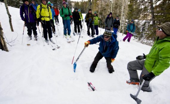 crowd of skiers stands and looks on as avalanche safety instructor digs in the snow to show snow layers