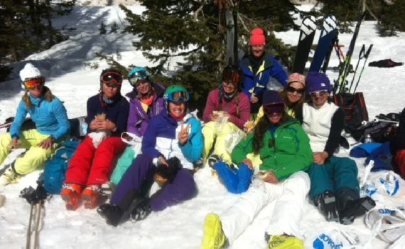 group of ski tester sitting in the snow in front of some pine trees, smiling for the photo and holding their homemade lunches