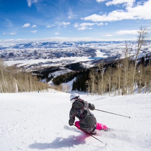 skier carves down a groomed Deer Valley run toward extensive mountain views on a sunny day