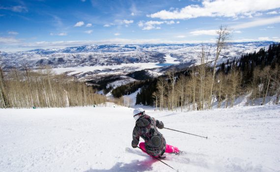 skier carves down a groomed Deer Valley run toward extensive mountain views on a sunny day