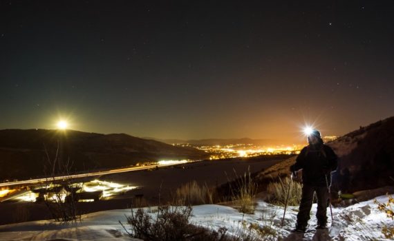 snowshoer wearing a headlamp looks out at the mountain view with city lights sparkling in the distance
