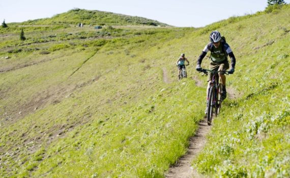 two mountain bikers ride along a cross-mountain trail, coming toward the photographer