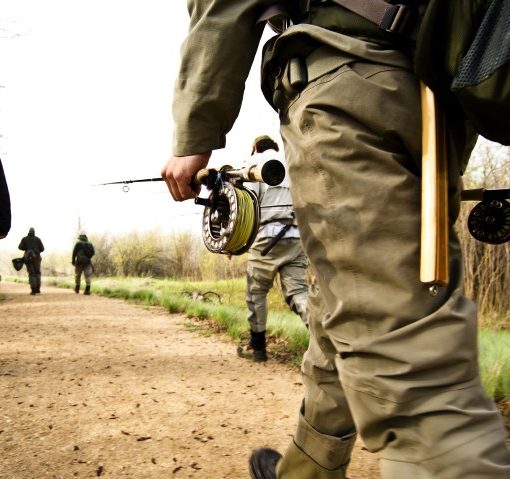 close up of one anglers fly rod in hand as multiple anglers walk along a dirt path toward the river