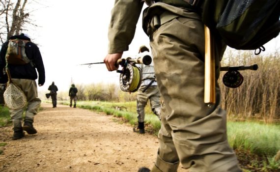 close up of one anglers fly rod in hand as multiple anglers walk along a dirt path toward the river