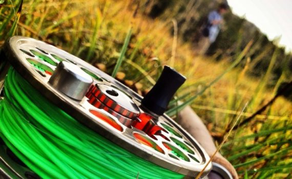 close-up view of a fly reel with a field and mountains in the background