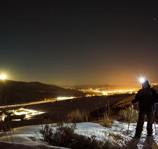 nighttime scene of snowshoer with a headlamp looking out over the town lights in Park City, Utah