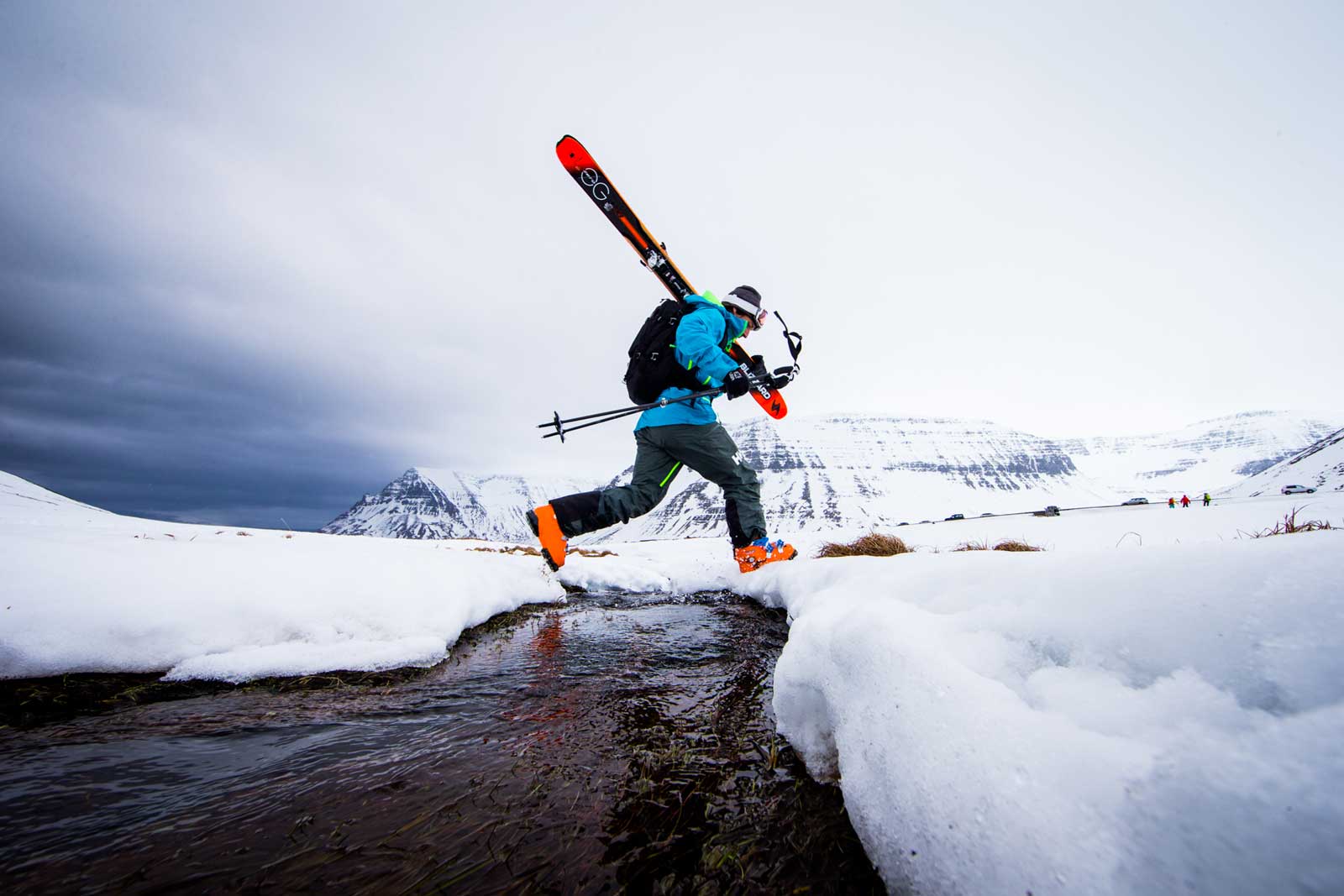 A skier jumps a river with skis on his shoulder