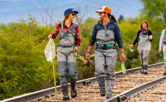 Four women with fly fishing gear walk down a rail road track near a river in Northern Utah