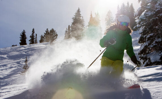 A woman skiing through deep powder with snow-covered trees in the background, her face beaming with joy as the sun shines brightly behind her. The golden light casts a hopeful glow, symbolizing a bright future for the outdoor industry.