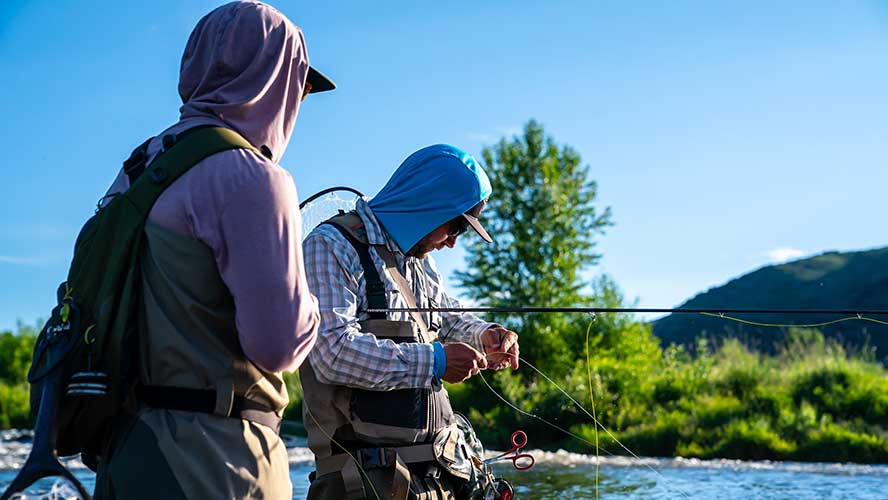 A guide and client stand in the river while the guide adjust his client's fishing line.