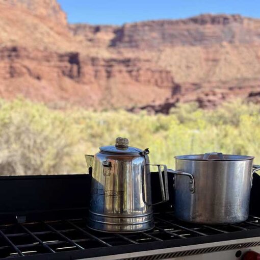 A camping stove is set up with a pot and kettle with desert landscape in the background.