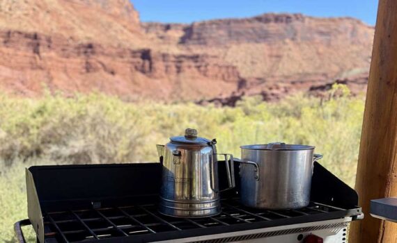 A camping stove is set up with a pot and kettle with desert landscape in the background.