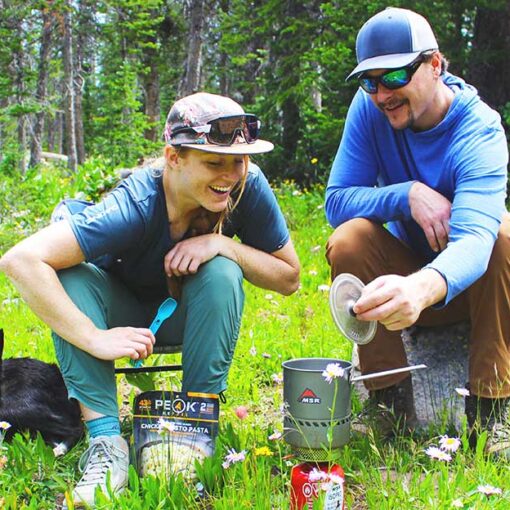 Jans Experts smile at their backcountry stove and see that their food is almost ready while their dog sits aside.