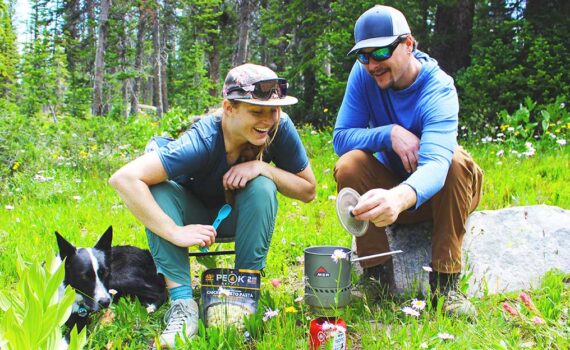 Jans Experts smile at their backcountry stove and see that their food is almost ready while their dog sits aside.