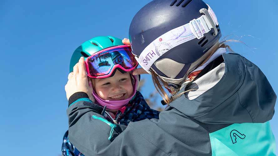 _MG_7275_888x500 Jans Expert adjusts the goggles on a young girl who is smiling on the ski hill.