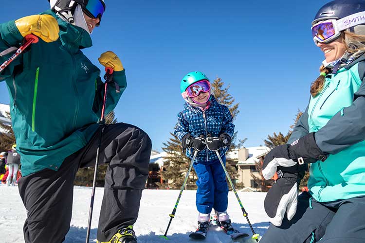 Child smiles on skis with parents on both sides of her in their ski rentals